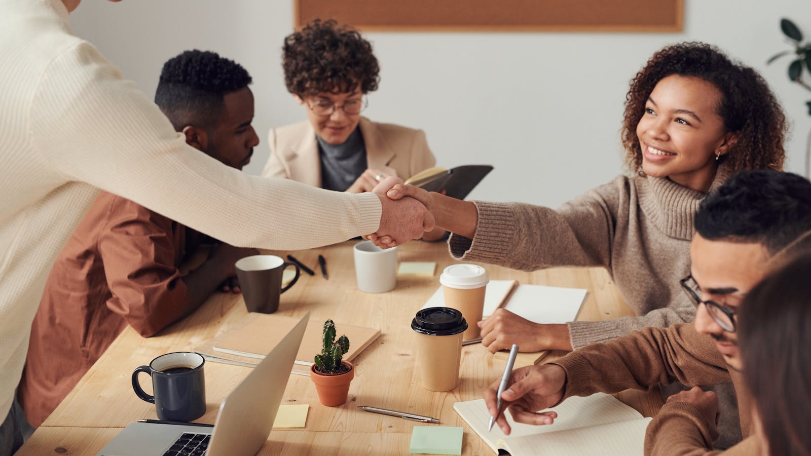 Colleagues collaborating indoors, sharing smiles, and shaking hands during a business meeting. This image shows diversity on product teams.